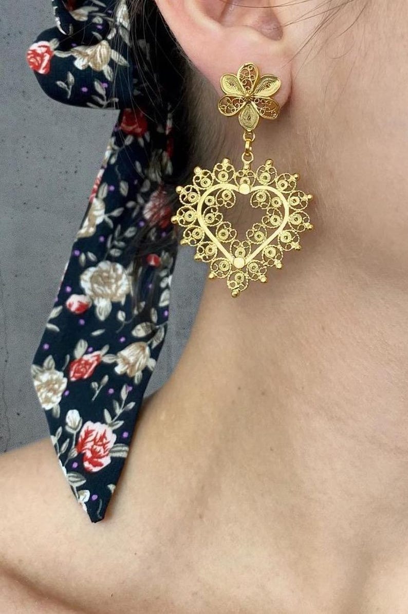 May include: Gold-toned filigree heart-shaped earrings with a floral accent. The earrings feature intricate openwork designs and a delicate, lacy appearance. A dark blue floral scarf is visible in the background.