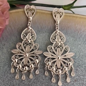 May include: A pair of silver filigree earrings. Each earring features a heart-shaped top, a floral design, and dangling teardrop-shaped charms. The intricate openwork design is visible against a soft background.