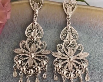 May include: A pair of silver filigree earrings. Each earring features a heart-shaped top, a floral design, and dangling teardrop-shaped charms. The intricate openwork design is visible against a soft background.