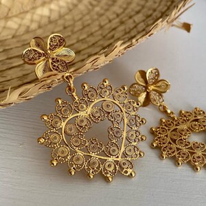 May include: Gold-colored filigree earrings. Each earring features a flower stud and a heart-shaped pendant with intricate openwork. The earrings are displayed on a light-colored surface, showcasing their delicate design.