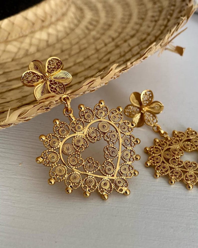 May include: Gold-colored filigree earrings. Each earring features a flower stud and a heart-shaped pendant with intricate openwork. The earrings are displayed on a light-colored surface, showcasing their delicate design.