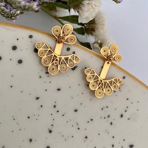May include: A pair of gold-tone earrings with a delicate filigree design. The earrings feature a fan-shaped design with a small flower detail at the top.