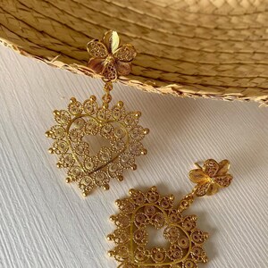 May include: A pair of gold-colored filigree earrings. Each earring features a flower-shaped stud, a delicate chain, and a heart-shaped pendant with intricate openwork. The earrings are displayed on a white surface.