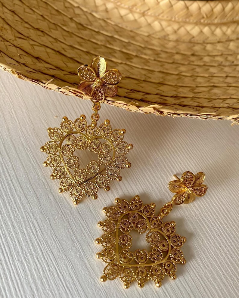 May include: A pair of gold-colored filigree earrings. Each earring features a flower-shaped stud, a delicate chain, and a heart-shaped pendant with intricate openwork. The earrings are displayed on a white surface.