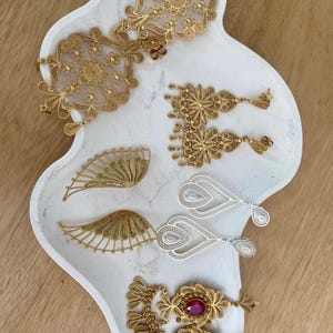May include: An assortment of filigree jewelry pieces in gold and silver tones, including earrings, pendants, and brooches, displayed on a white, organically shaped tray. The jewelry features intricate floral and geometric designs, with some pieces incorporating red gemstones.