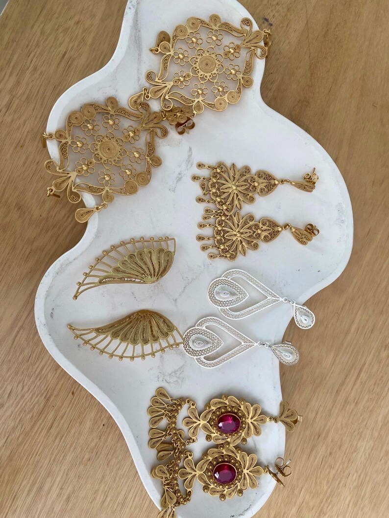 May include: An assortment of filigree jewelry pieces in gold and silver tones, including earrings, pendants, and brooches, displayed on a white, organically shaped tray. The jewelry features intricate floral and geometric designs, with some pieces incorporating red gemstones.
