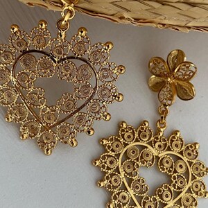 May include: Gold-toned filigree earrings. One earring features a flower stud, while the other has a heart-shaped pendant. The intricate design includes circular and floral patterns, creating a delicate and ornate appearance. The earrings are likely for pierced ears.