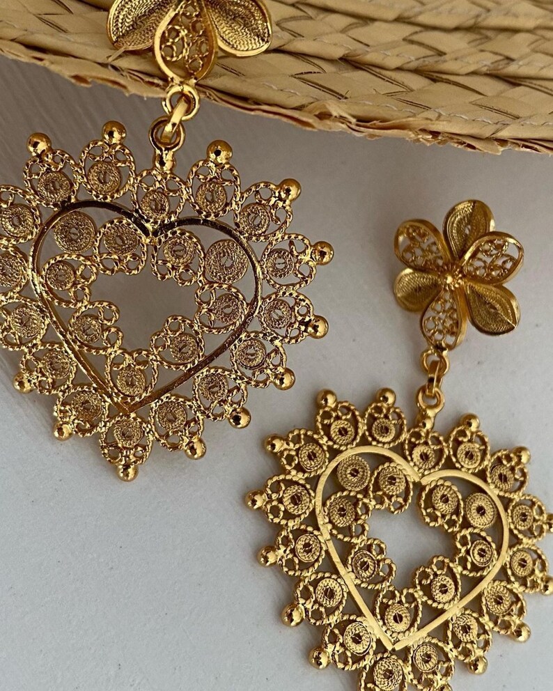 May include: Gold-toned filigree earrings. One earring features a flower stud, while the other has a heart-shaped pendant. The intricate design includes circular and floral patterns, creating a delicate and ornate appearance. The earrings are likely for pierced ears.