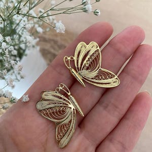 May include: Two gold-colored butterfly-shaped earrings. The earrings are made of a delicate filigree design. The butterfly wings have intricate patterns. The earrings are displayed on a hand, with white flowers in the background.