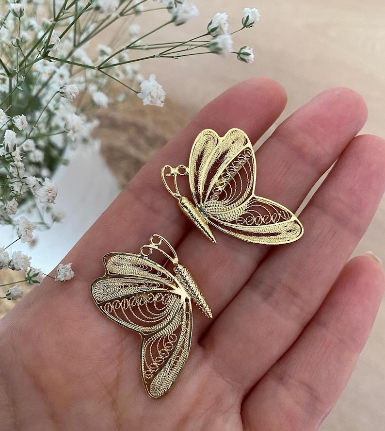 May include: Two gold-colored butterfly-shaped earrings. The earrings are made of a delicate filigree design. The butterfly wings have intricate patterns. The earrings are displayed on a hand, with white flowers in the background.