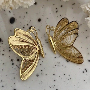 May include: Two gold-colored butterfly-shaped brooches. The brooches have intricate filigree detailing on the wings and a textured body. The butterflies are displayed on a speckled white surface.