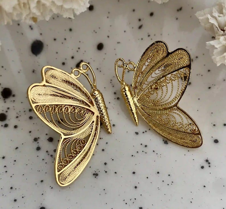 May include: Two gold-colored butterfly-shaped brooches. The brooches have intricate filigree detailing on the wings and a textured body. The butterflies are displayed on a speckled white surface.