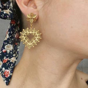 May include: Gold filigree earrings. The earrings feature a flower stud and a heart-shaped drop. The heart-shaped drop is intricately designed with delicate filigree work. The earrings are a warm gold color and are worn on the ear.