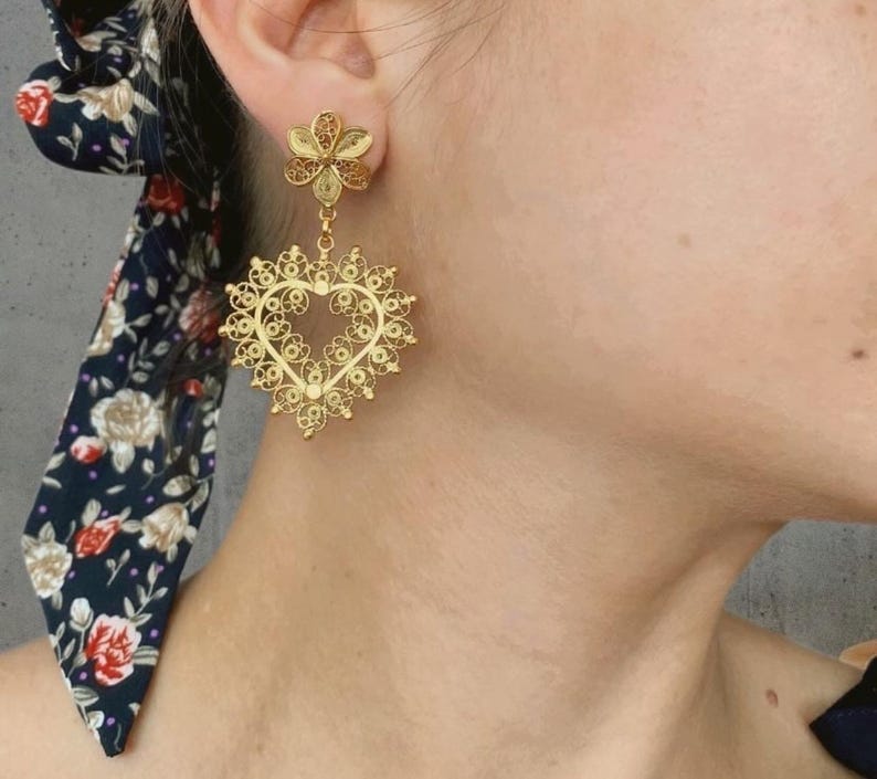 May include: Gold filigree earrings. The earrings feature a flower stud and a heart-shaped drop. The heart-shaped drop is intricately designed with delicate filigree work. The earrings are a warm gold color and are worn on the ear.
