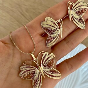 May include: A gold-colored jewelry set featuring a butterfly pendant necklace and matching butterfly earrings. The butterfly designs have intricate filigree detailing. The jewelry is displayed on a hand.