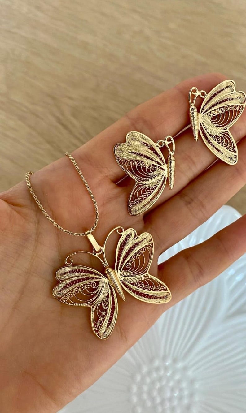 May include: A gold-colored jewelry set featuring a butterfly pendant necklace and matching butterfly earrings. The butterfly designs have intricate filigree detailing. The jewelry is displayed on a hand.