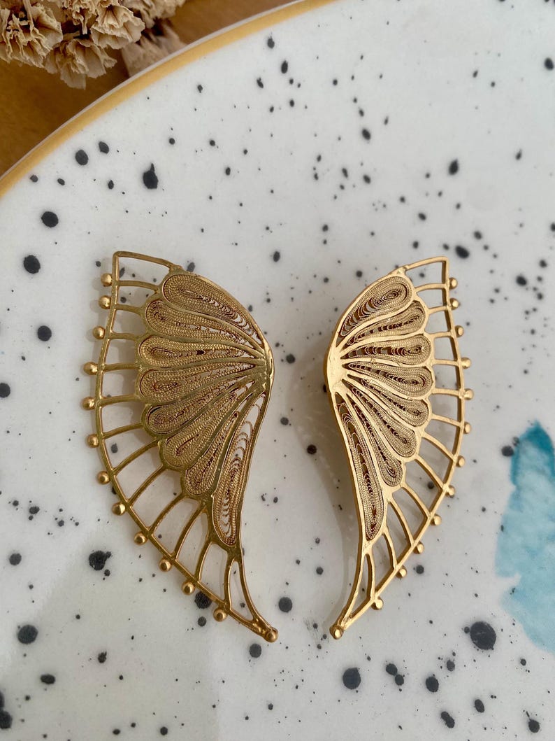 May include: A pair of gold-colored filigree butterfly wing earrings. The earrings are displayed on a speckled white and gray ceramic plate. The wings have intricate detailing and a delicate, open design.