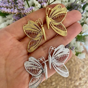 May include: Close-up of three filigree butterfly pendants in gold and silver. The butterflies have intricate, openwork designs. The gold butterflies are a warm, bright yellow, while the silver butterflies have a cool, metallic sheen. The pendants are displayed against a soft background.