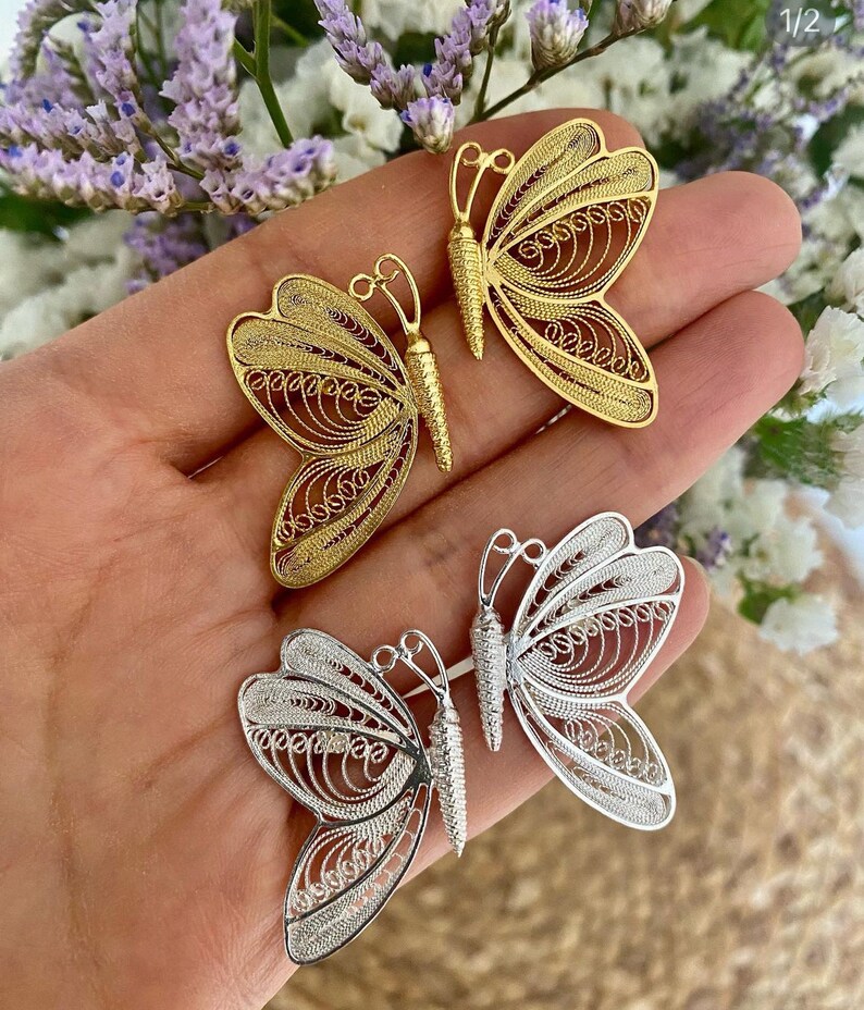 May include: Close-up of three filigree butterfly pendants in gold and silver. The butterflies have intricate, openwork designs. The gold butterflies are a warm, bright yellow, while the silver butterflies have a cool, metallic sheen. The pendants are displayed against a soft background.