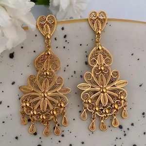 May include: A pair of gold filigree earrings with a heart-shaped top and a flower-shaped bottom. The earrings have a dangling design with small beads and teardrop-shaped accents.