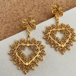 May include: A pair of gold-colored filigree earrings. Each earring features a flower-shaped stud, connected to a heart-shaped pendant with an open center. The heart is surrounded by a detailed, ornate design. The earrings are displayed on a white surface.