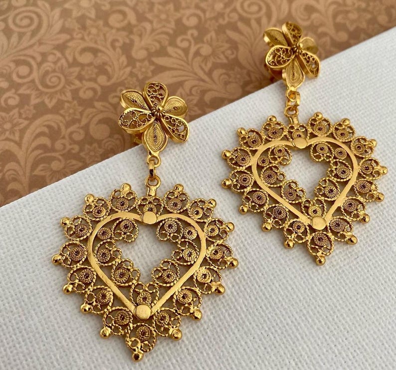 May include: A pair of gold-colored filigree earrings. Each earring features a flower-shaped stud, connected to a heart-shaped pendant with an open center. The heart is surrounded by a detailed, ornate design. The earrings are displayed on a white surface.
