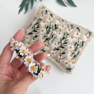 Op de afbeelding: Een beige haarband met witte en gele madeliefjesaccenten wordt voor een beige kussen met een madeliefjespatroon gehouden. De haarband heeft een kanten rand en het kussen heeft een crèmekleurige rand. De madeliefjes hebben gele centra en groene stelen.