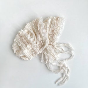 May include: A white lace bonnet with a ruffled edge and long ties. The bonnet is made of delicate fabric and has a vintage look.