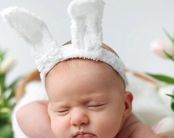May include: A sleeping baby wearing a white bunny headband with soft, fluffy ears. The baby is lying on a white surface with a blurred background of flowers.