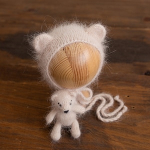 May include: A white knitted baby hat with fuzzy ears and a matching white knitted teddy bear. The hat is on a wooden form and the bear is sitting on a wooden surface.