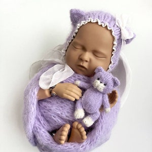 Puede incluir: Una muñeca de bebé de punto morado con un gorro de encaje blanco y un juguete de gato de punto morado. La muñeca está envuelta en una manta de punto morado.