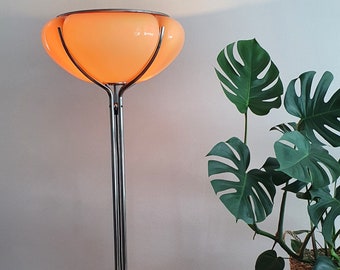 Puede incluir: Una lámpara de pie de cromo con una pantalla grande, redonda y de color naranja. La lámpara está sobre un suelo de madera junto a una planta en maceta y una mesita auxiliar de madera.