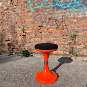 May include: A bright orange and black stool with a round, black padded seat. The stool has a unique, flared base and a silver band around the center.