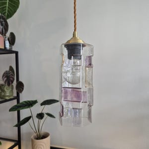 May include: A rectangular glass pendant light with a brass fitting and a brown cord. The light fixture is clear with sections of lavender and clear glass. An exposed lightbulb is visible inside. The light hangs against a white wall.