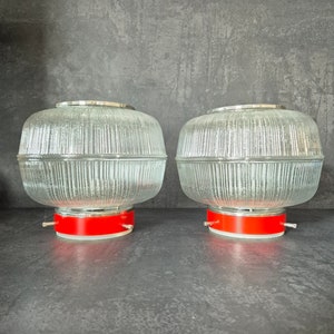 May include: Two clear glass lampshades with a ribbed design and red bases. The lampshades are cylindrical and have a silver trim around the top.