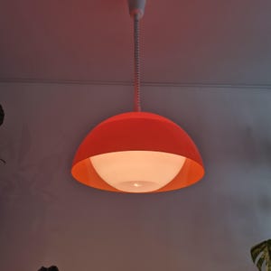 Könnte beinhalten: Eine moderne orangefarbene Pendelleuchte mit weißem Innenraum. Das Licht hängt an einem weißen Kabel und ist an der Decke befestigt. Die Leuchte hat eine abgerundete, schalenförmige Form.
