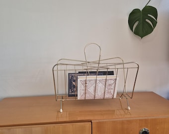 May include: A gold metal magazine rack with a white handle and four legs. The rack is holding three books, including one titled "Great Goddesses".