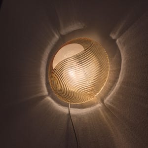 Op de afbeelding: Een ronde, amberkleurige wandlamp met een textuur, golvend ontwerp. De lamp straalt een warm licht uit en werpt schaduwen op de muur. De lamp is op een witte muur gemonteerd en heeft een zichtbaar netsnoer.