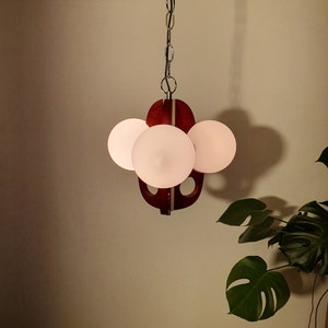 May include: A contemporary pendant light with four spherical, frosted glass bulbs. The fixture has a dark wood and metal design, suspended from a silver chain. The light hangs against a neutral wall, with a large green plant in the corner, creating a stylish interior.