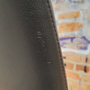 以下が含まれることがあります： 黒い革の椅子背もたれのアップ。背もたれは縫い付けられた縫い目で湾曲しており、滑らかで質感のある表面をしています。