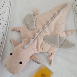 Può includere: Un peluche a forma di drago rosa con dettagli grigi, tra cui ali e una spina dorsale. Ha occhi neri a bottone ed è accompagnato da un piccolo impacco di gel giallo.