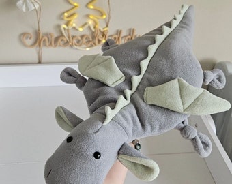 May include: A gray plush dragon toy with light green accents on the wings and spine. The dragon has black button eyes and is being held up by a person's hand. The background includes a white surface and a decorative sign that says "Baby".