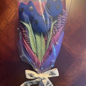 May include: A bouquet of crocheted blue flowers with green stems and purple crocheted leaves wrapped in clear plastic with a white ribbon with a bee pattern.