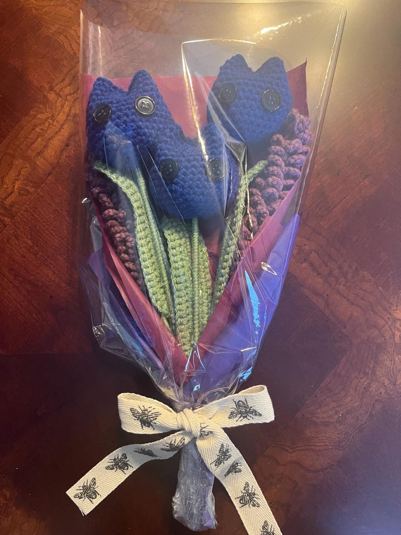 May include: A bouquet of crocheted blue flowers with green stems and purple crocheted leaves wrapped in clear plastic with a white ribbon with a bee pattern.