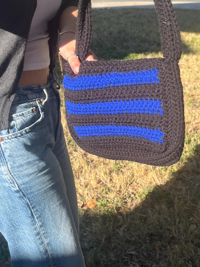 May include: A black and blue crocheted crossbody bag with a striped pattern. The bag has a long strap and is made of yarn.