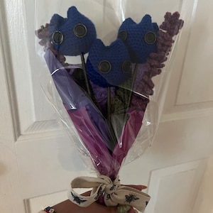 May include: A bouquet of blue crocheted flowers with button eyes, wrapped in purple and clear plastic with a white ribbon tied around the stems.