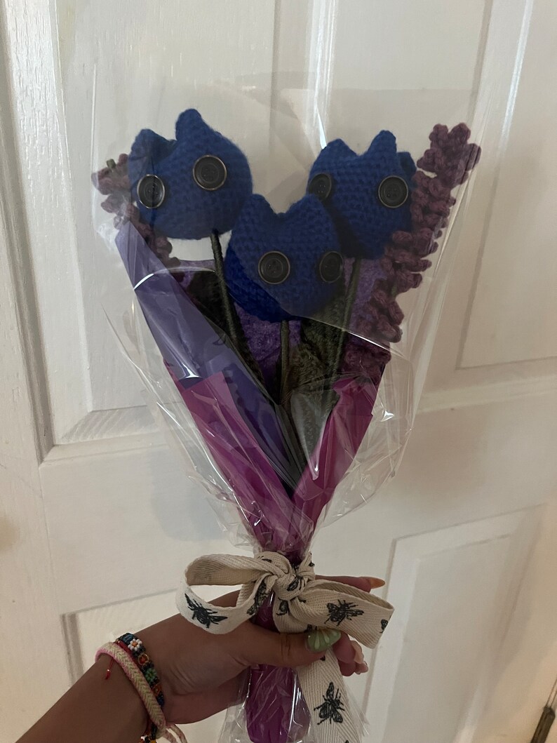 May include: A bouquet of blue crocheted flowers with button eyes, wrapped in purple and clear plastic with a white ribbon tied around the stems.