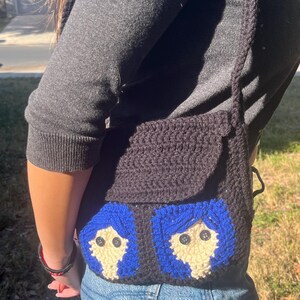May include: A black crocheted crossbody bag with two blue and white faces on the front. The faces have blue hair, white skin, and black button eyes.
