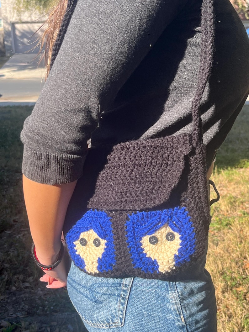 May include: A black crocheted crossbody bag with two blue and white faces on the front. The faces have blue hair, white skin, and black button eyes.