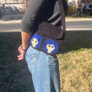 May include: A black crocheted shoulder bag with two blue and white embroidered faces. The bag has a long strap.